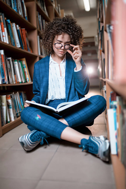Młoda piękna kręcone dziewczyna w okularach i niebieski garnitur siedzący z książkami w bibliotece. - Zdjęcie, obraz