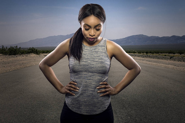 Athletic black African American female runner or jogger resting to pose with a motivated facial expression with a road during sunset in the background.  Depicts endurance and determination during marathon sports.  - Foto, imagen