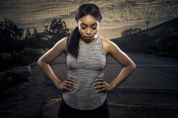 Atletische zwarte African American vrouwelijke runner of jogger rusten om te poseren met een gemotiveerde gezichtsuitdrukking met een weg tijdens zonsondergang op de achtergrond. Verbeeldt uithoudingsvermogen en vastberadenheid tijdens Marathon sporten.  - Foto, afbeelding