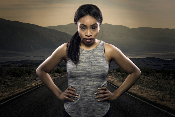 Athletic black African American female runner or jogger resting to pose with a motivated facial expression with a road during sunset in the background.  Depicts endurance and determination during marathon sports.  - Foto, Imagen