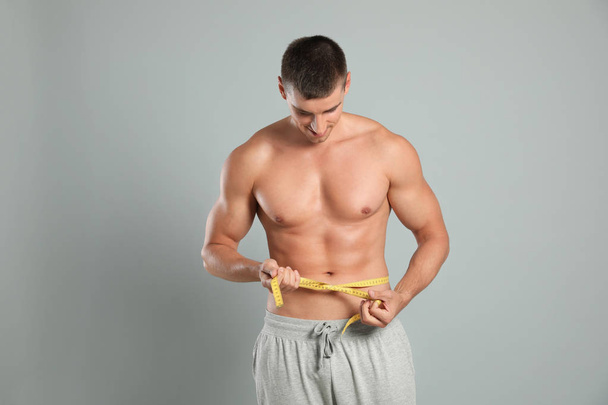 Young man with slim body using measuring tape on grey background - Foto, Imagem