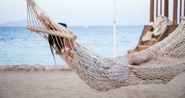 Nuori nainen istuu riippumatossa meren rannalla ja rentoutuu
. - Valokuva, kuva