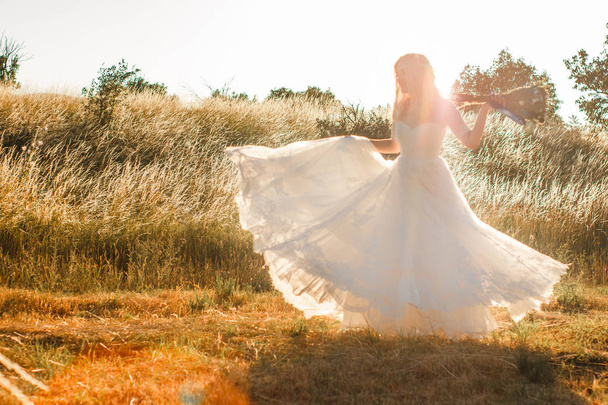 Bride on a summer field in white wedding dress rolling and dancing in sunset light. Sun beams seen through transparent dress skirt fabric. Rustic or boho outdoor wedding concept. Selective soft focus. - Fotó, kép
