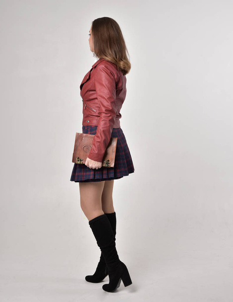 Ganzkörperporträt eines brünetten Mädchens in roter Lederjacke und kariertem Rock, das stehend mit einem Buch auf cremefarbenem Studiohintergrund posiert. - Foto, Bild
