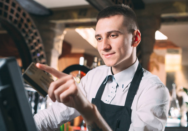 small business, people and service concept - happy man or waiter in apron at counter with cashbox working at bar or coffee shop. - Foto, Bild