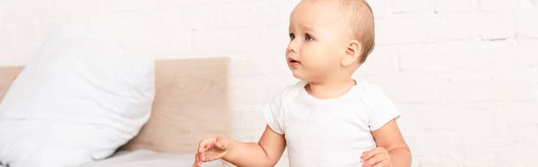 Panoramic shot of cute little child in white bodysuit sitting near wooden bed with pillow - Photo, Image