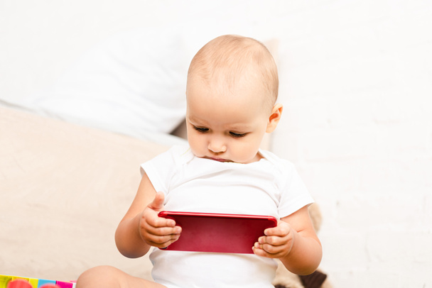 Cute chubby kid looking at red smartphone in white room - Foto, Imagem