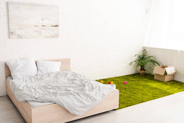 Sala de luz com cama de casal e caixa de papelão perto da planta no tapete verde
 - Foto, Imagem