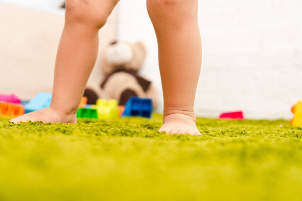Vista recortada de un niño descalzo parado en el suelo verde entre juguetes coloridos
 - Foto, Imagen