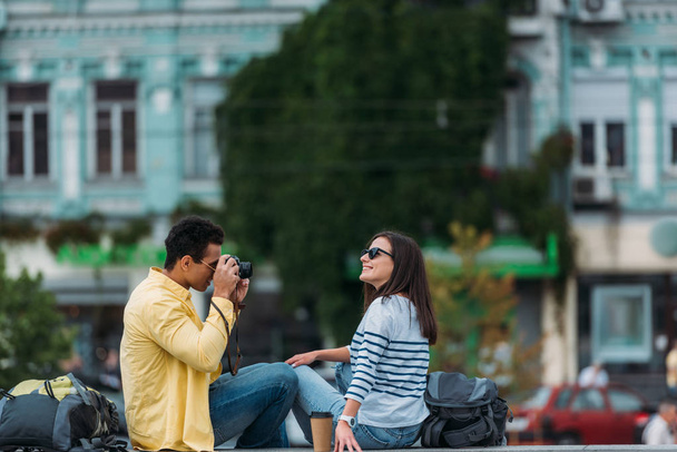 Двурасовый мужчина сидит и фотографирует женщину в солнечных очках с рюкзаком
 - Фото, изображение