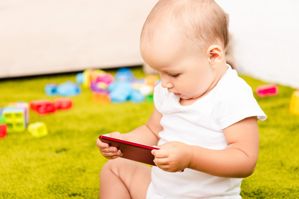 niedliches kleines Kind sitzt auf grünem Boden mit Spielzeug und hält digitales Gerät - Foto, Bild