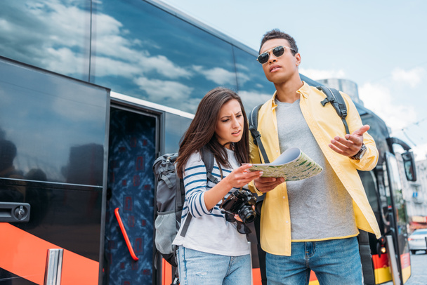 Mujer con cámara digital mirando hacia arriba en el mapa con el hombre de raza mixta cerca de autobús de viaje
 - Foto, imagen