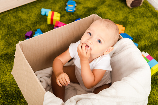 Cute blue-eyed child sitting in cardboard box with white blanket on green floor - Foto, Imagem