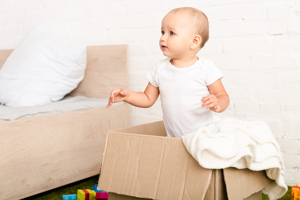 Little child standing in cardboard box with white blanket and looking away - Foto, immagini