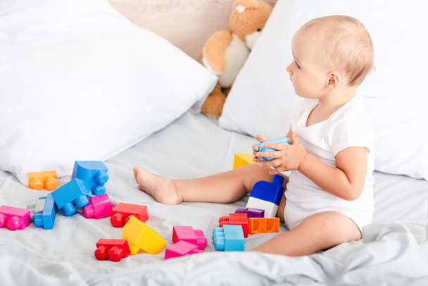 Barefoot little child in white clothes sitting on bed with pillows near colorful toys  - Photo, Image