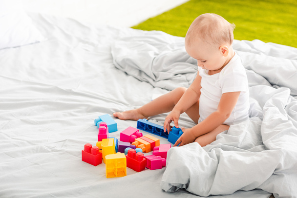 cute barefoot baby in white clothes sitting on bed and playing with colorful toys - Φωτογραφία, εικόνα