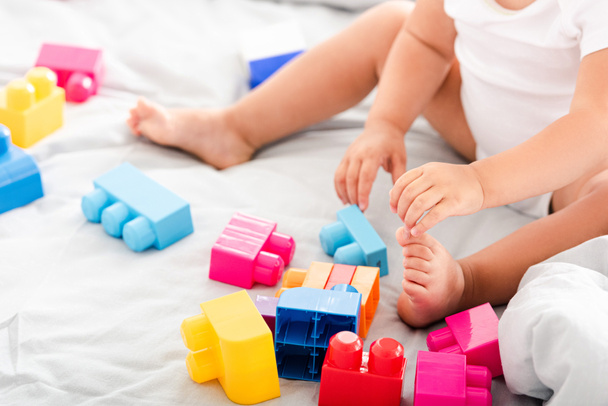 Partial view of barefoot baby in white clothes sitting on bed and playing with bright construction - Foto, immagini