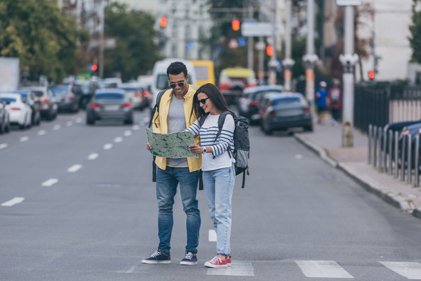 Woman and multiracial friend with backpacks holding city map on crosswalk - Фото, зображення