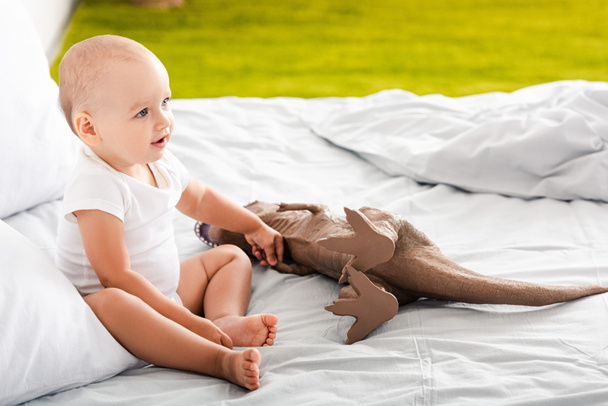 Cute barefoot child in white clothes smiling near brown dinosaur toy - Photo, Image
