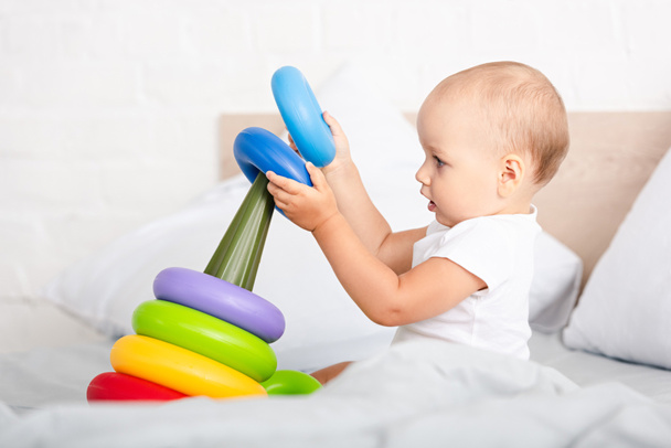 Side view of cute little child in white clothes playing with toy pyramid on bed - Foto, imagen