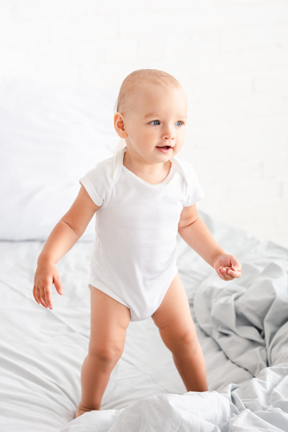 Cute little child in white clothes standing on bed, smiling and looking away - Foto, Bild