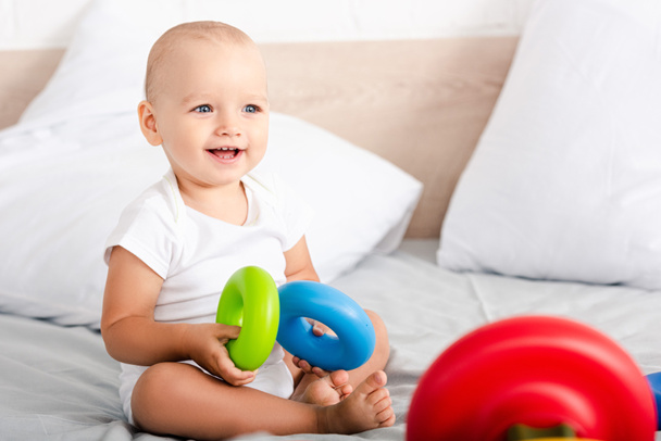 Criança bonito com sorriso sentado na cama e segurando anéis de brinquedo coloridos
 - Foto, Imagem