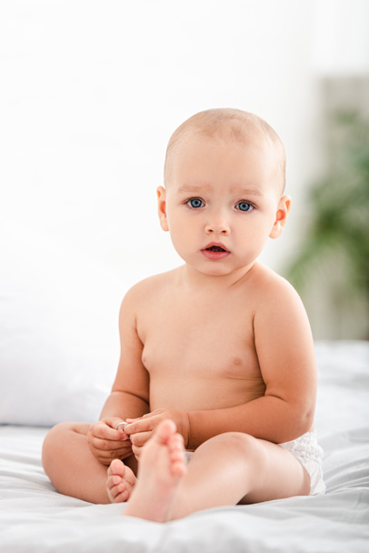 Cute barefoot child with blue eyes sitting on bed and looking at camera - Foto, imagen