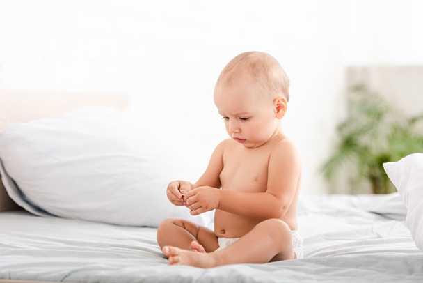Cute barefoot child in diaper sitting on bed in ligth room - Foto, Imagen