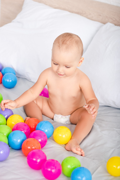 Top view of cute little baby sitting on bed among colored balls - Foto, Imagem