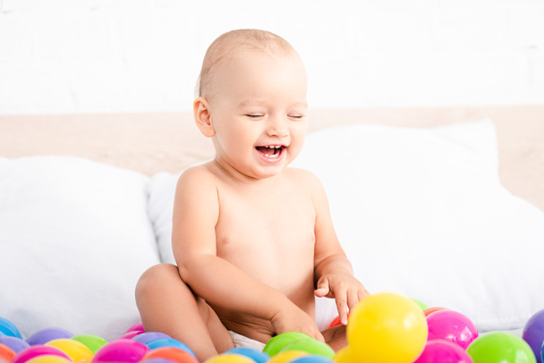 Cute little baby sitting on bed, playing with colored balls and laughing with closed eyes - Fotografie, Obrázek