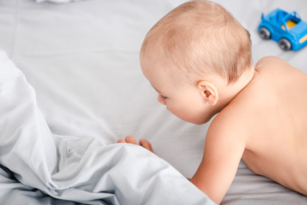 Вид сзади маленького ребенка, лежащего на кровати рядом с игрушечной машиной
 - Фото, изображение
