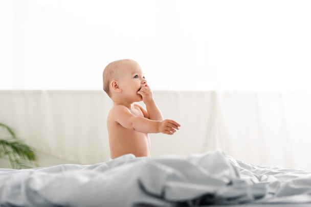 Vue latérale du mignon petit enfant assis sur le lit et prenant les mains dans la bouche
 - Photo, image