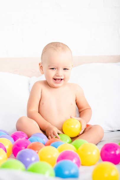 Cute little baby sitting on bed, playing with colored balls and laughing - Photo, Image