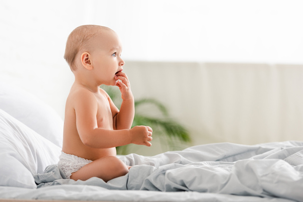 Vista lateral del bebé mirando hacia otro lado y tomando los dedos en la boca
 - Foto, Imagen
