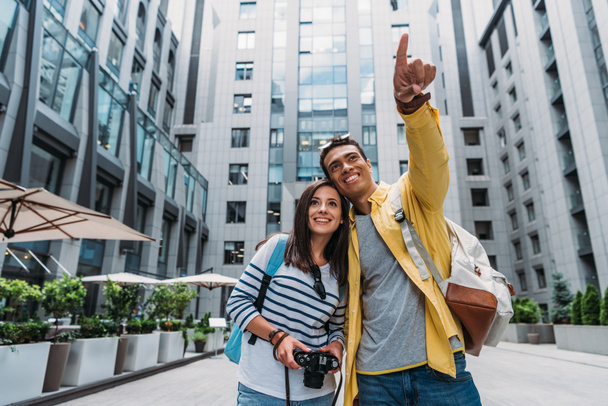 happy girl holding digital camera near cheerful mixed race man pointing with finger  - 写真・画像