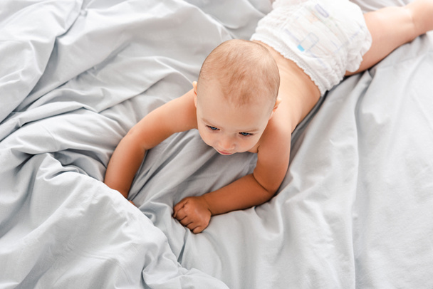 Cute baby in diaper laying on white bedding and looking away - Photo, Image