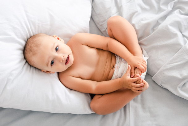 Top uitzicht van kleine baby in luier leggen op rug en grijpen voeten met handen en kijken naar camera - Foto, afbeelding