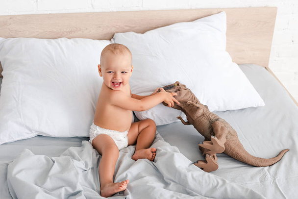 Ευτυχισμένο μικρό παιδί κάθεται στο κρεβάτι, χαμόγελο και παίζοντας με καφέ παιχνίδι δεινόσαυρος - Φωτογραφία, εικόνα