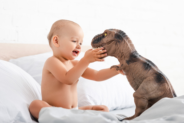 Niño pequeño sentado en la cama blanca y sosteniendo dinosaurio juguete con ambas manos
 - Foto, imagen