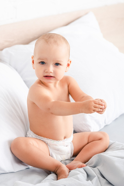 Cute blue-eyed child sitting on bed with white bedclothes and looking at camera - Photo, Image