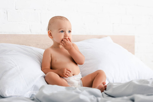 Mezítláb kis gyermek ül az ágyban, és figyelembe ujjak szájába - Fotó, kép