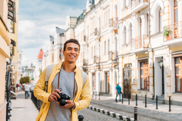 cheerful mixed race man smiling while holding digital camera near buildings  - 写真・画像