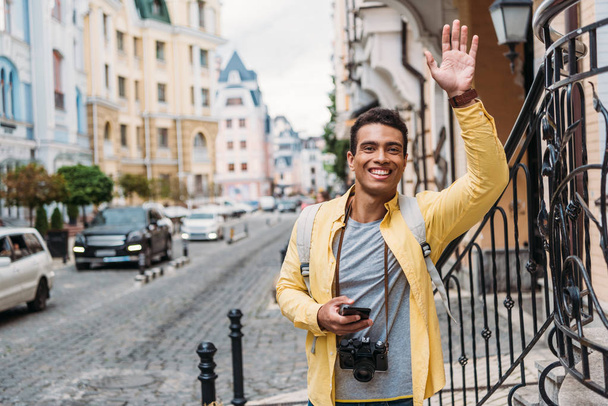 happy mixed race man holding smartphone while waving hand  - Foto, afbeelding