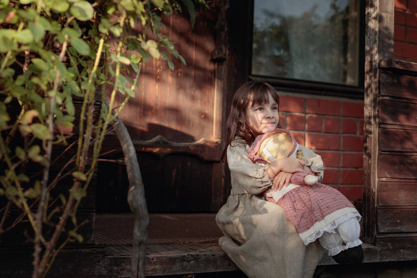 Κορίτσι με χειροποίητη κούκλα σε ένα καλοκαιρινό βράδυ κοντά στο παλιό σπίτι. - Φωτογραφία, εικόνα