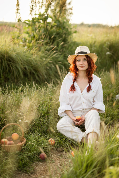 Ένα νεαρό κορίτσι με ένα ψάθινο καπέλο που κρατά ένα ροδάκινο και κάθεται σε ένα μονοπάτι - Φωτογραφία, εικόνα