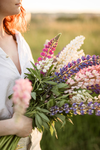 A red hair woman holding a bouquet of lupine, lupin flowers during sunset hour - Photo, Image