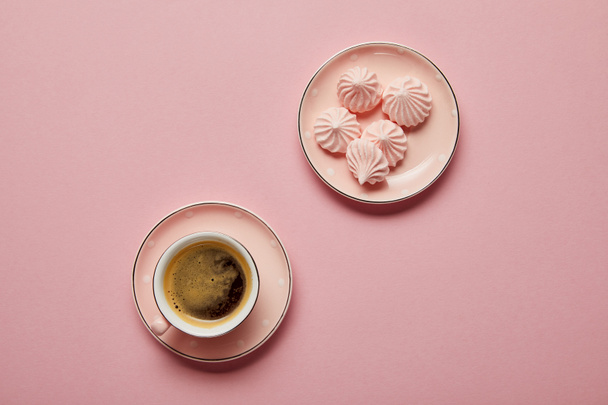Top view of small pink meringues on dotted saucer and cup of coffee on pink background - Photo, Image