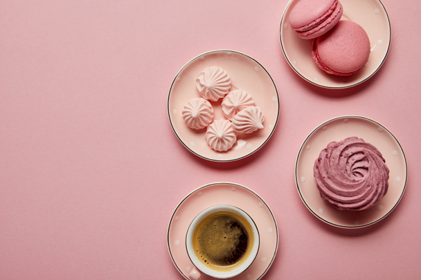 Top view of pink meringues and macaroons on small pink saucers with white dots and cup of coffee on pink background - 写真・画像