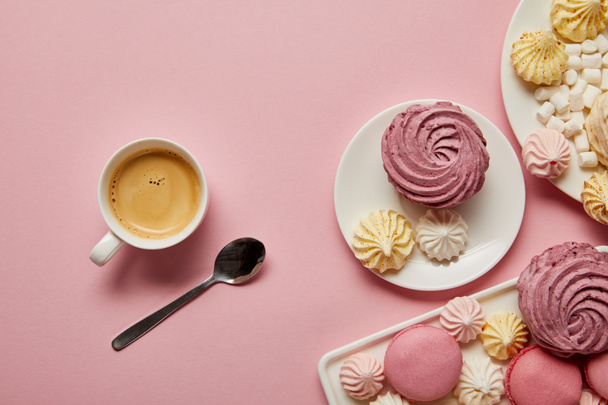 Top view of pink, white and yellow meringues, macaroons, marshmallows and cup of coffee with spoon on pink background - Fotografie, Obrázek