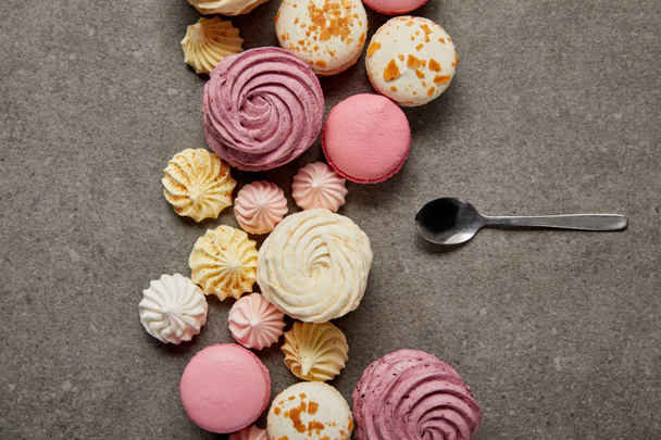 Top view of pink and white macaroons with pink, yellow and white meringues with teaspoon on gray background - Foto, Bild